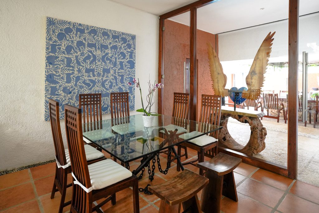 Comedor con mesa de cristal, sillas talladas y mural de azulejo azul en Casa Corazón